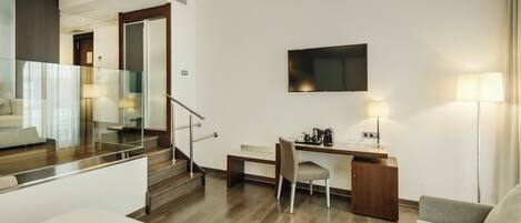 A modern hotel room with a wooden staircase, a flat-screen TV, a desk with a chair, a sofa, and a lamp.
