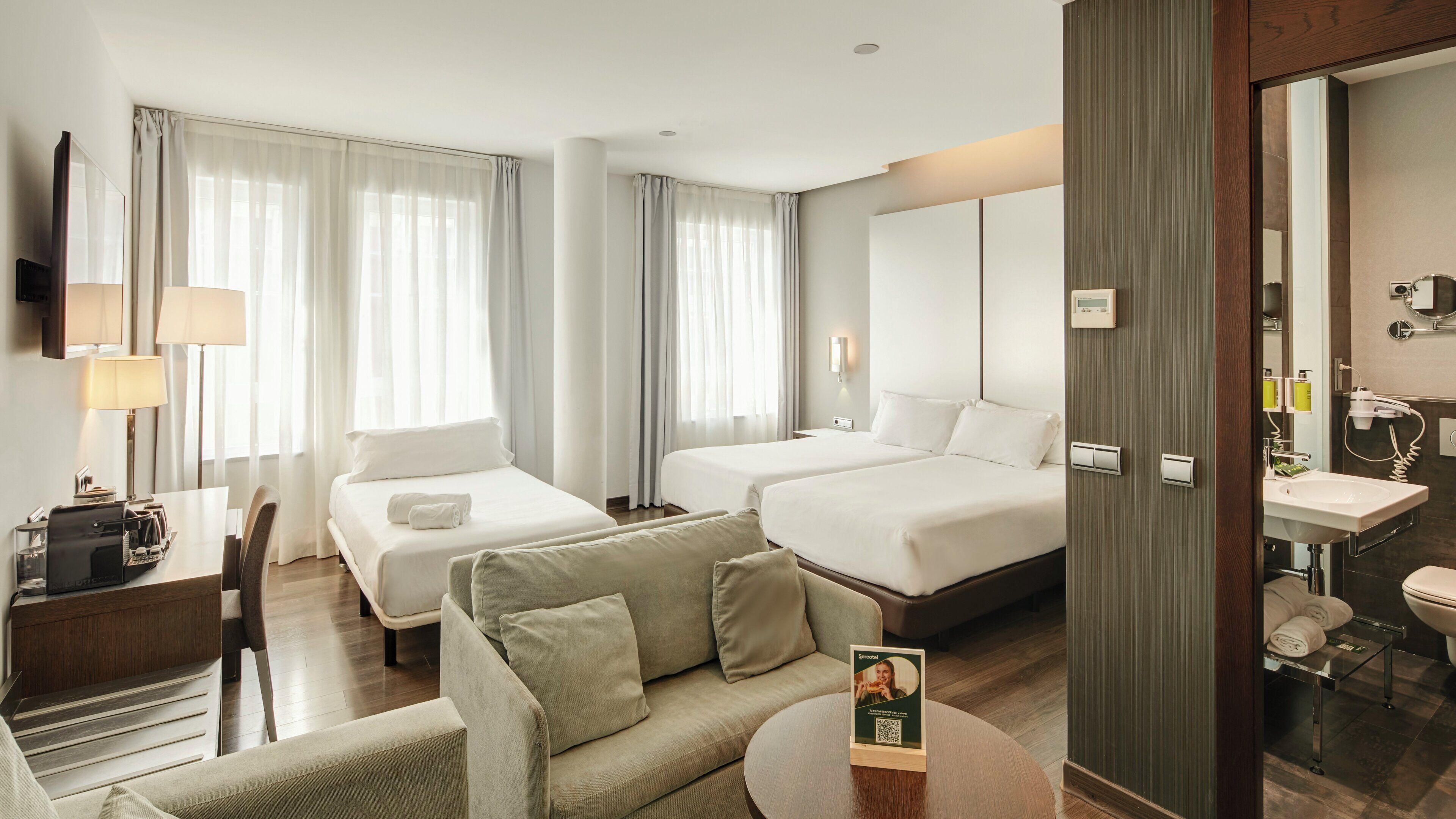Une chambre d’hôtel avec deux lits, un canapé, un bureau avec une lampe et une salle de bain équipée d’un lavabo et d’un miroir.
