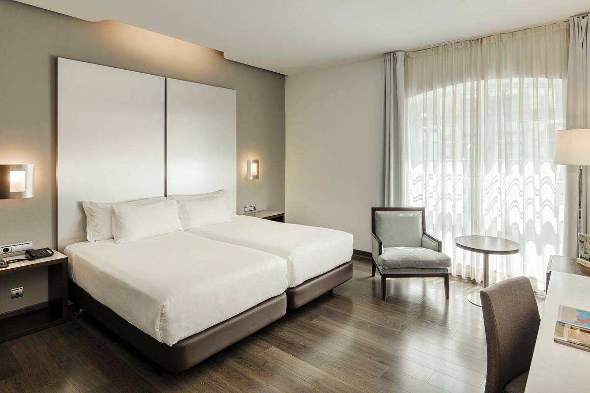 Ein Hotelzimmer mit Doppelbett, Nachttischen, einem Sessel, einem Schreibtisch und einem großen Fenster mit Vorhängen.