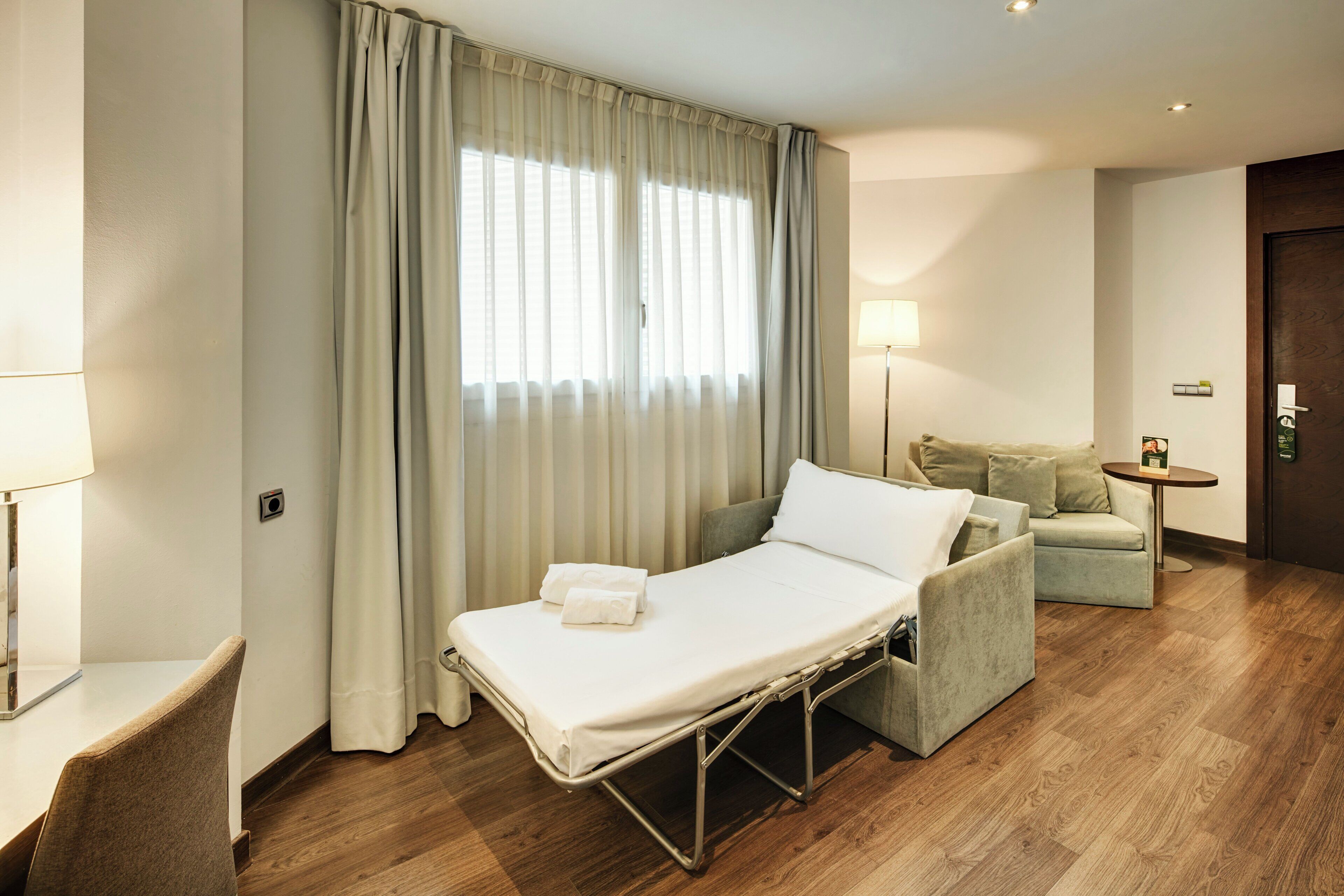 Habitación de hotel con una cama individual, una mesita de noche, una lámpara, un sofá y una pequeña mesa con otra lámpara.