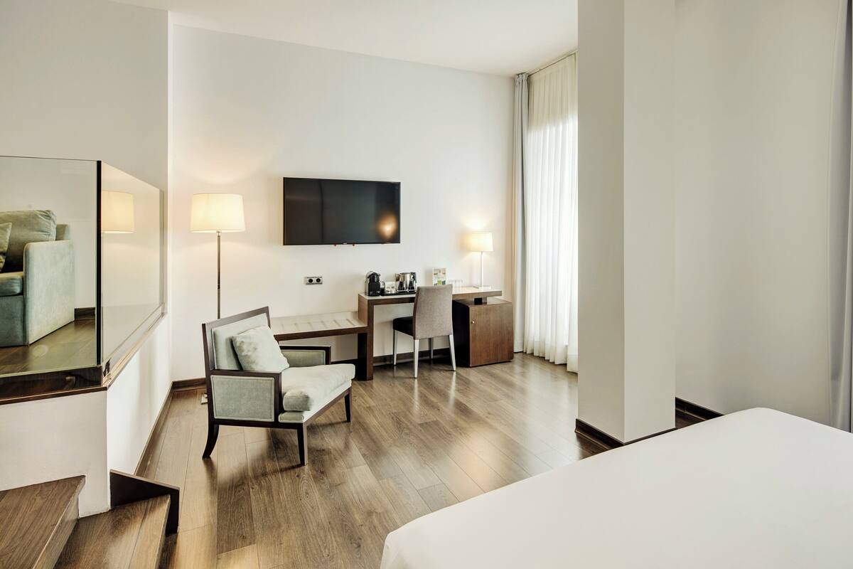 A hotel room with a bed, a desk, a chair, a TV, and a large window with curtains.