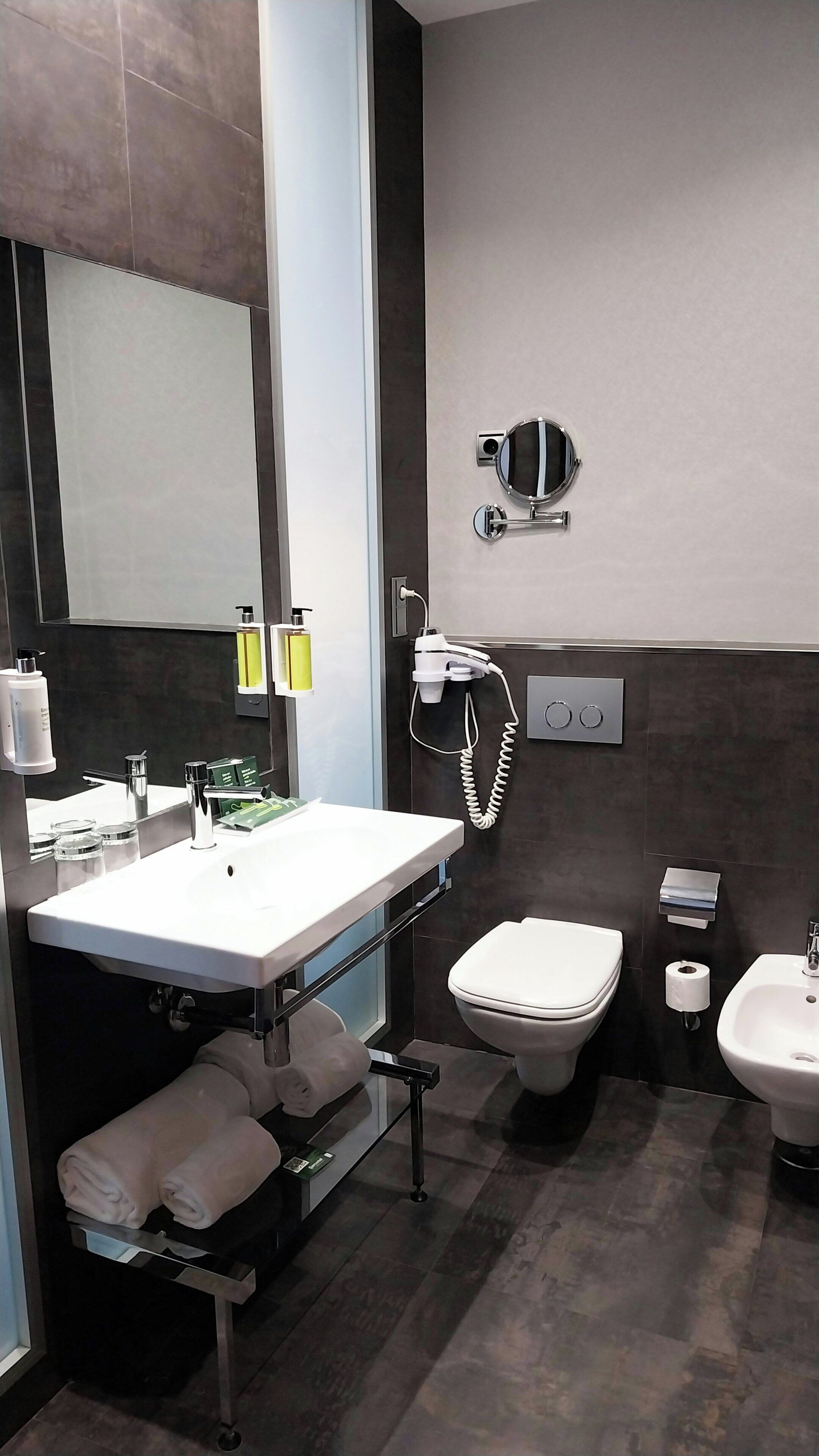 Une salle de bain moderne avec un grand miroir, un lavabo avec un meuble-vasque, des toilettes, un bidet et un sèche-cheveux fixé au mur.