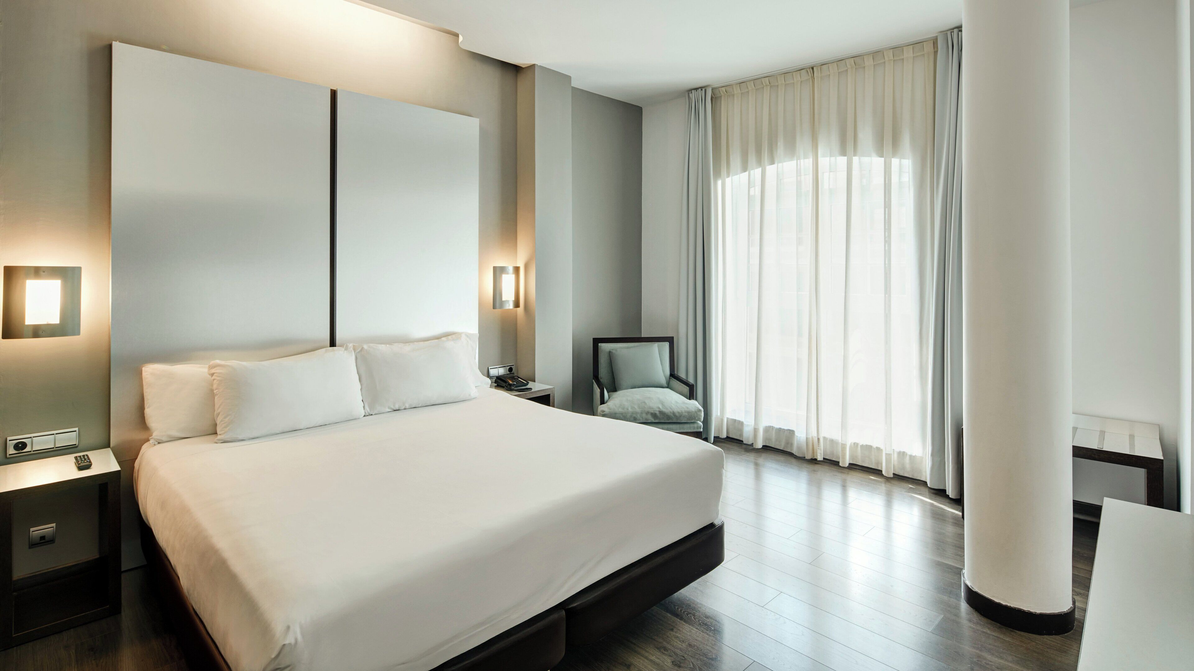 Una habitación de hotel moderna con una cama grande, lámparas de noche, una silla y un ventanal con cortinas.