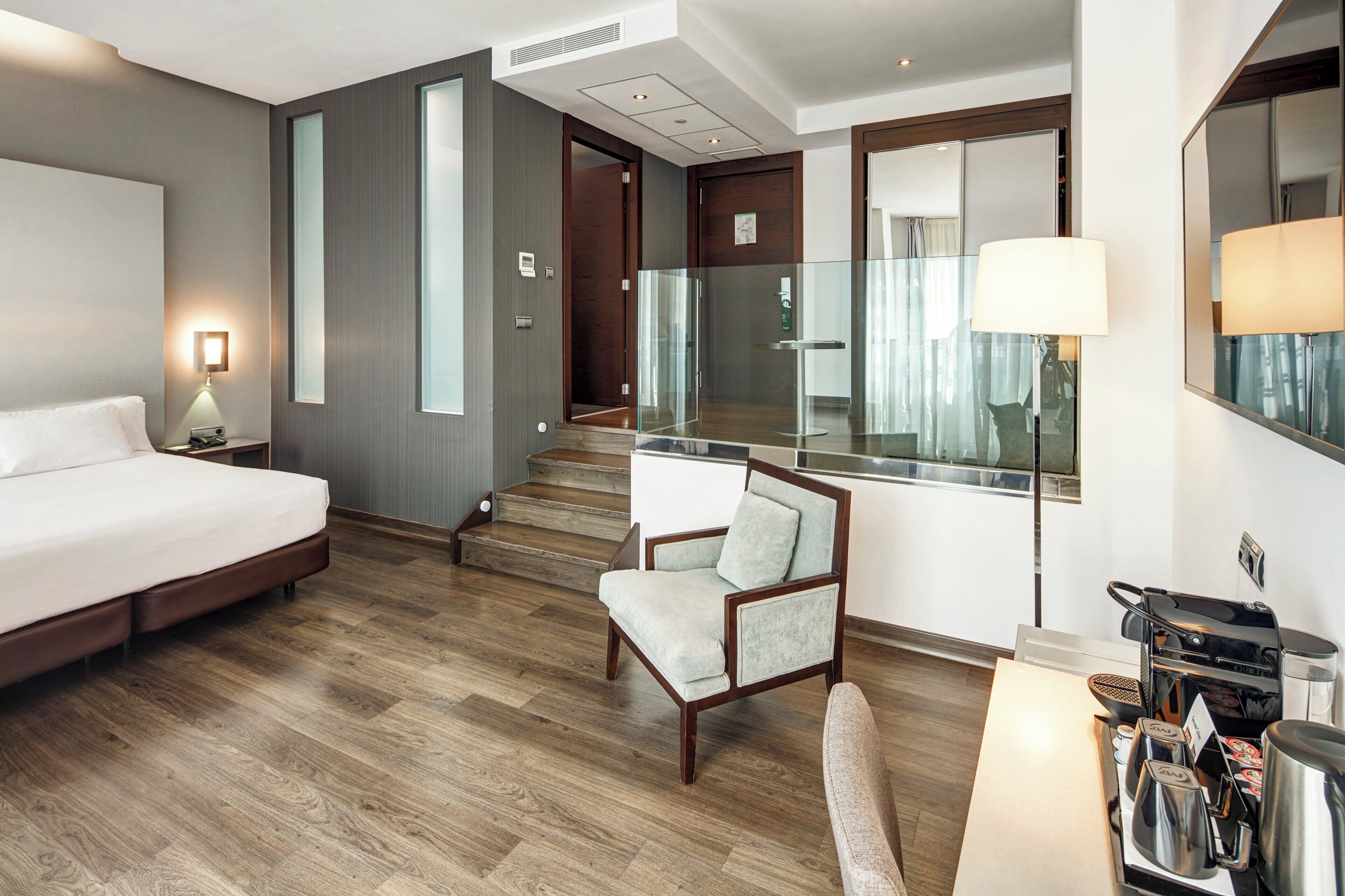 A hotel room with a bed, a chair, a desk with a coffee maker, and a staircase leading to an upper floor.