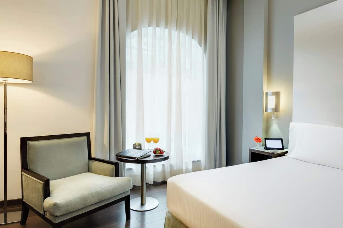Ein Hotelzimmer mit Bett, Sessel, kleinem Tisch mit Laptop, Lampe und einem Fenster mit Vorhängen.