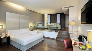 A modern hotel room with a large bed, a desk with a coffee maker, and a dining area with fruit and a glass of orange juice. - Hotel Sercotel Coliseo (Bilbao)