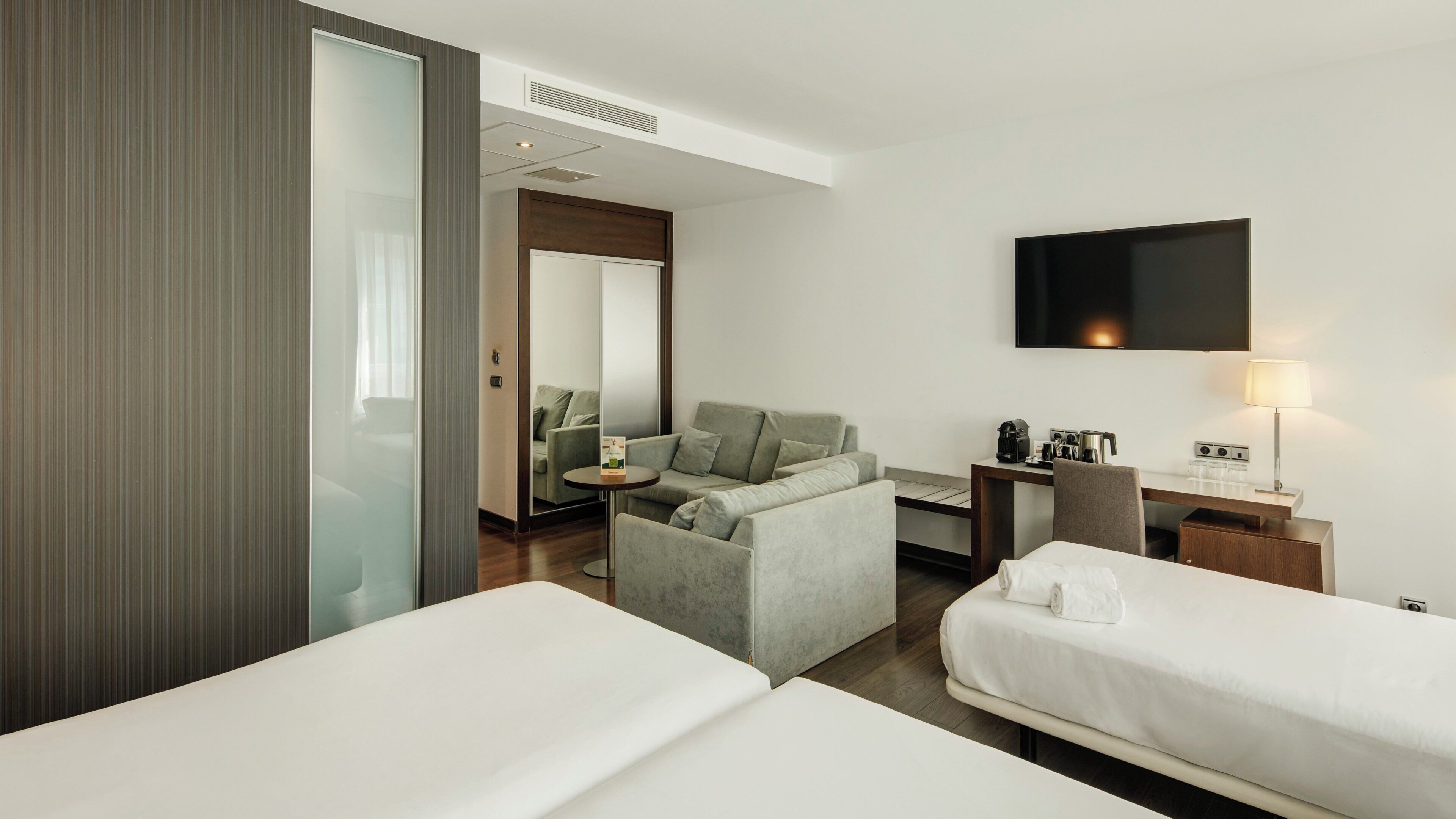 A hotel room with two beds, a sofa, a desk with a lamp, a TV, and a coffee maker.