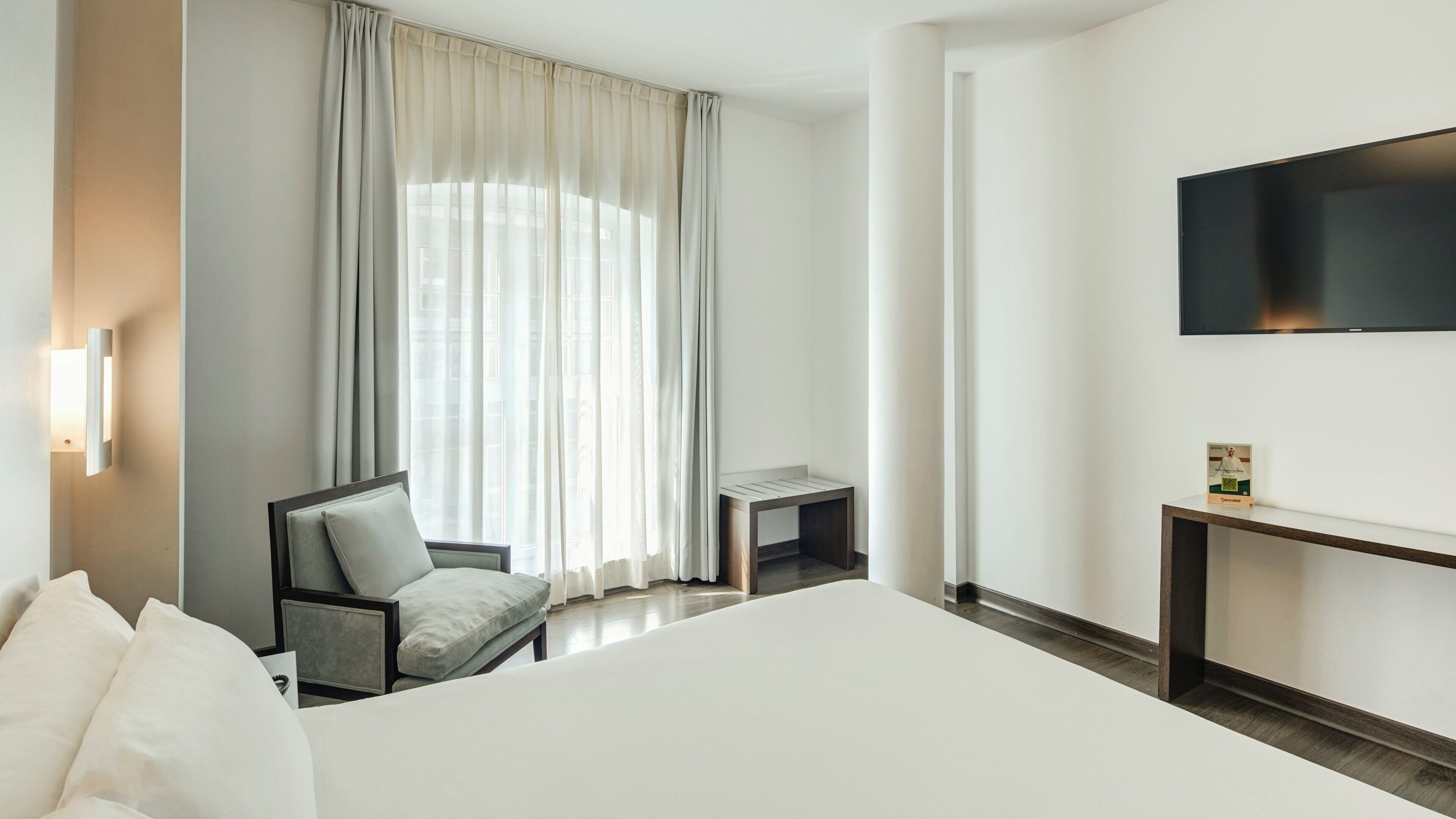 A hotel room with a bed, a chair, a small table, a TV, and a window with curtains.