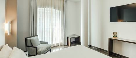 A hotel room with a bed, a chair, a small table, a TV, and a window with curtains.