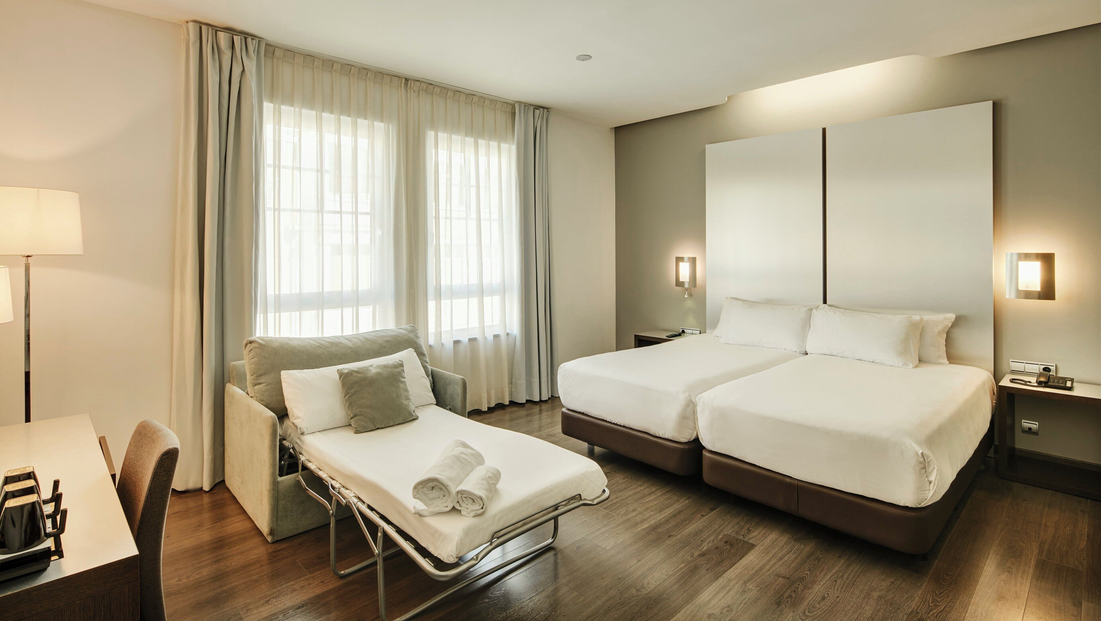 A hotel room with two beds, a desk, a chair, a bedside table, and a window with curtains.