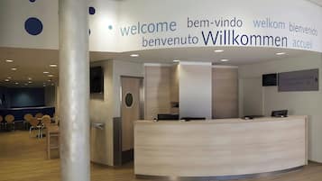 A modern reception area with a central desk, a seating area with chairs, and a sign with multilingual welcome messages.