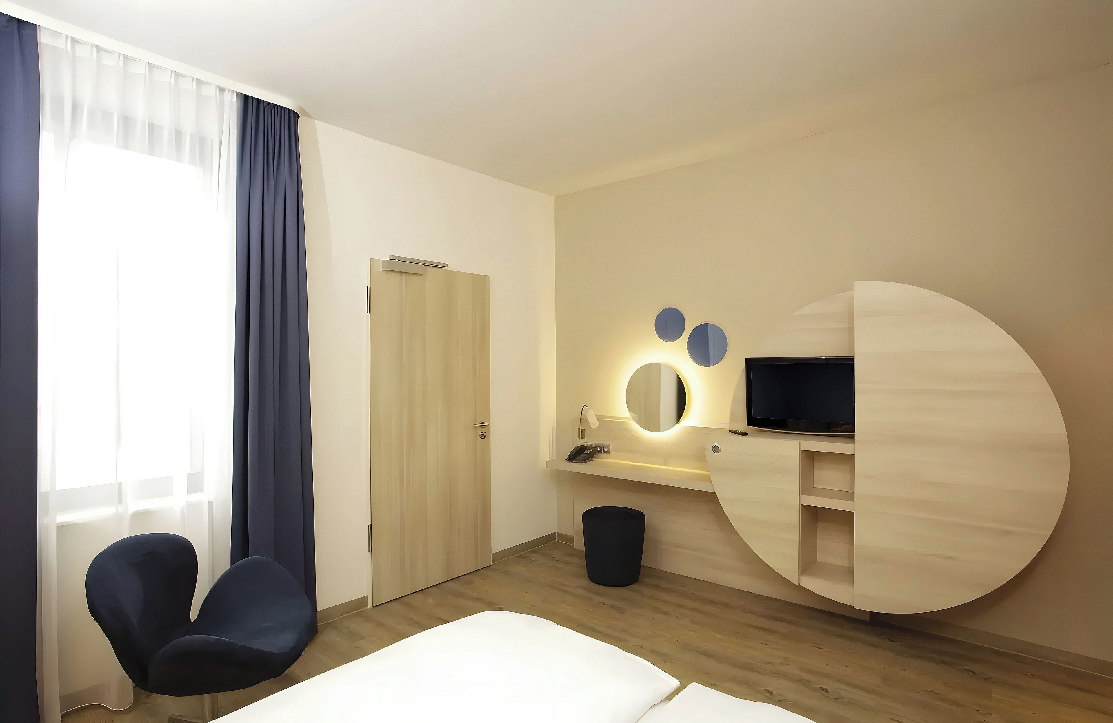 Ein Hotelzimmer mit einem Bett, einem Schreibtisch mit Flachbildfernseher, einem Sessel, einer Mülleimer und einer Tür.