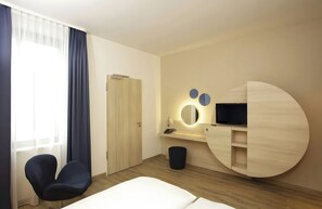 Una habitación de hotel con cama, escritorio con televisor de pantalla plana, una silla, un cesto de basura y una puerta.