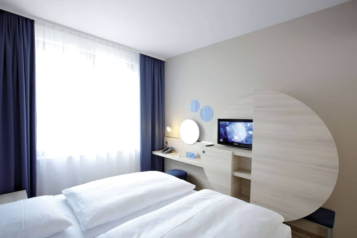 Una habitación de hotel moderna con una cama, un escritorio con computadora, un televisor y un ventanal con cortinas.
