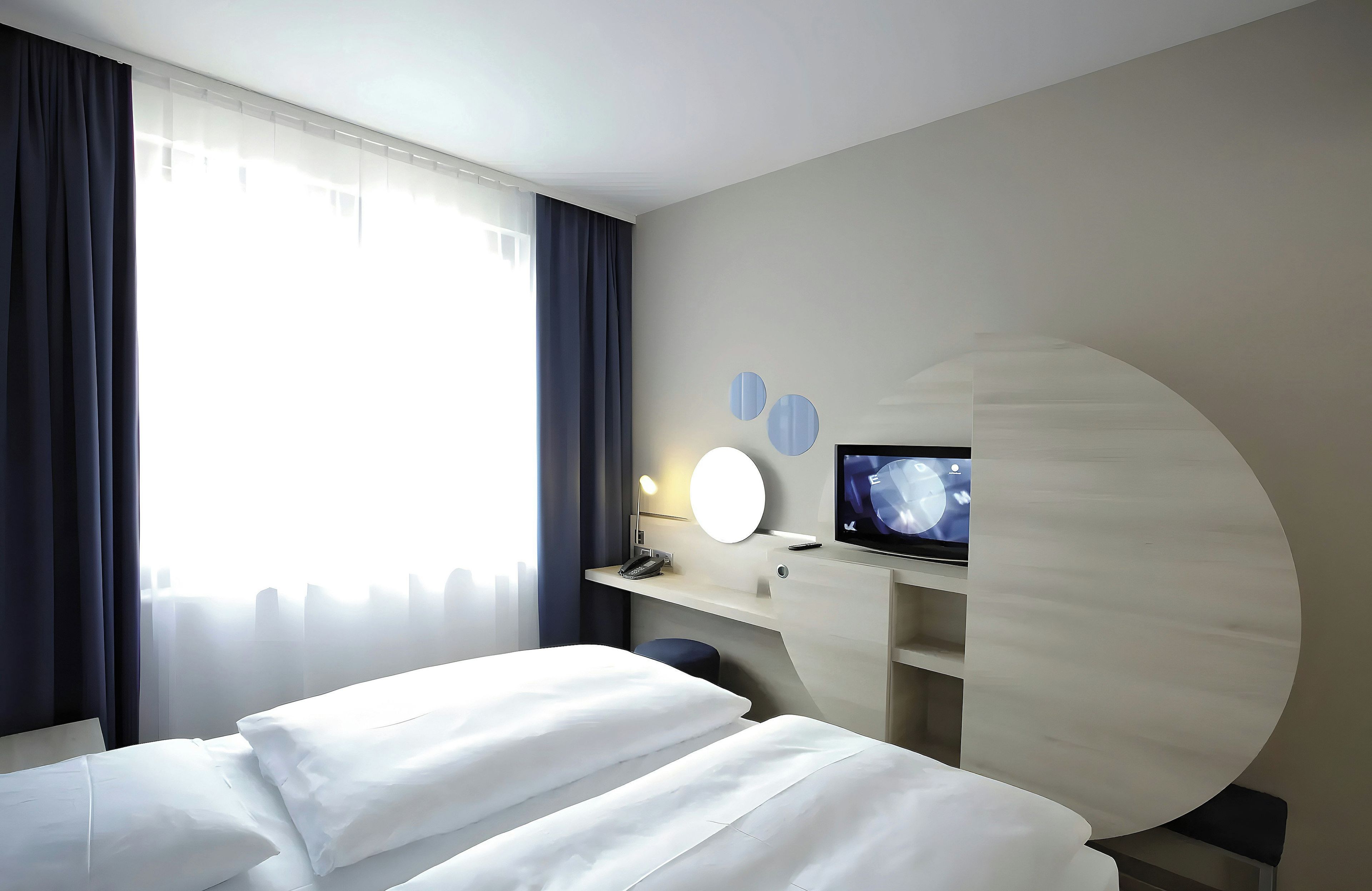 A hotel room with a bed, a desk with a computer monitor, and a window with curtains.