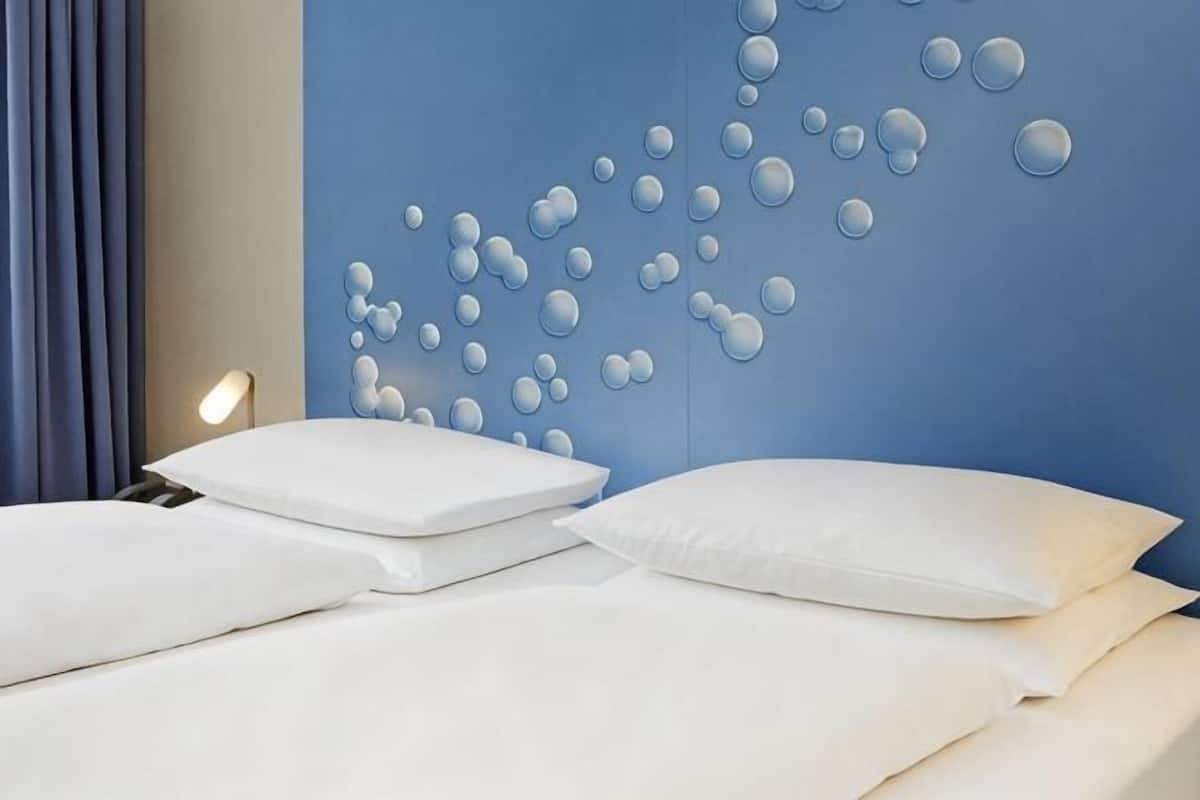 Habitación de hotel con una pared azul de acento, cama con ropa blanca y una lámpara de pared.