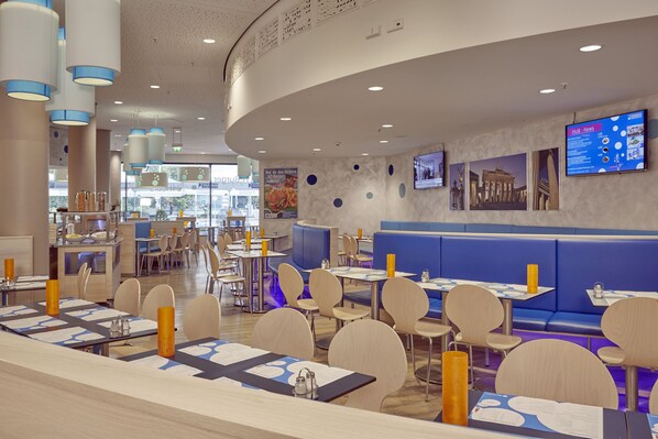 A modern restaurant with round tables, blue booths, and a digital menu board. -  Leonardo Smart Hotel Berlin City East  (Berlin)
