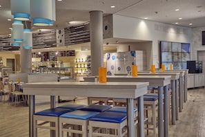 Relax in our spacious seating area with high tables and blue cushioned chairs, surrounded by a modern reception desk and a well-organized bar.