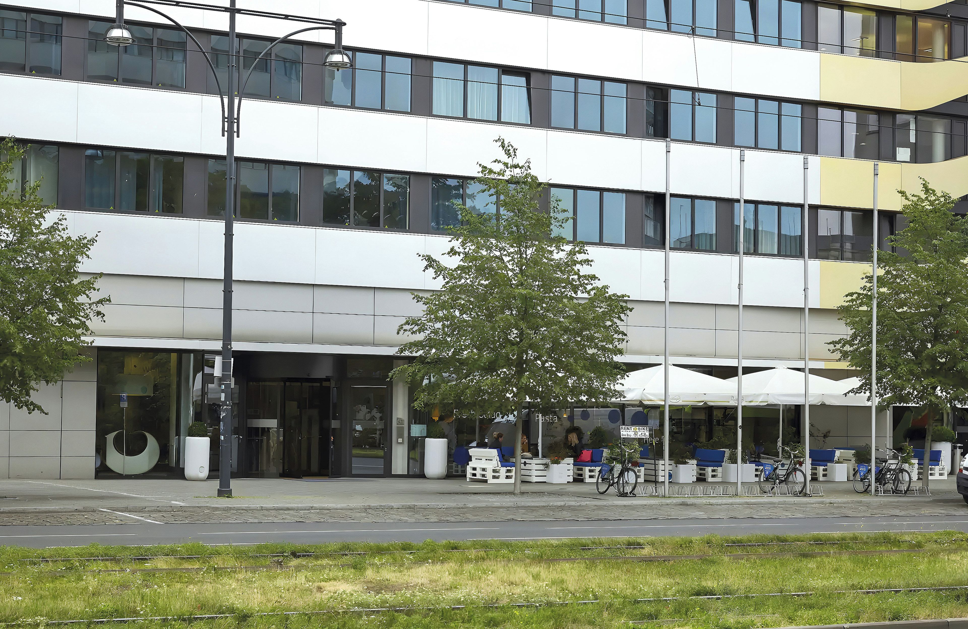 Un edificio moderno con fachada de vidrio, zona de estar al aire libre y un cartel de alquiler de bicicletas.