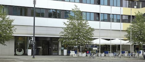 Un edificio moderno con fachada de vidrio, zona de estar al aire libre y un cartel de alquiler de bicicletas.