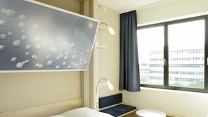 A modern hotel room with a bed, a desk, two chairs, a small table, and a window with a view of buildings. - Leonardo Smart Hotel Berlin Alexanderplatz (Berlin)
