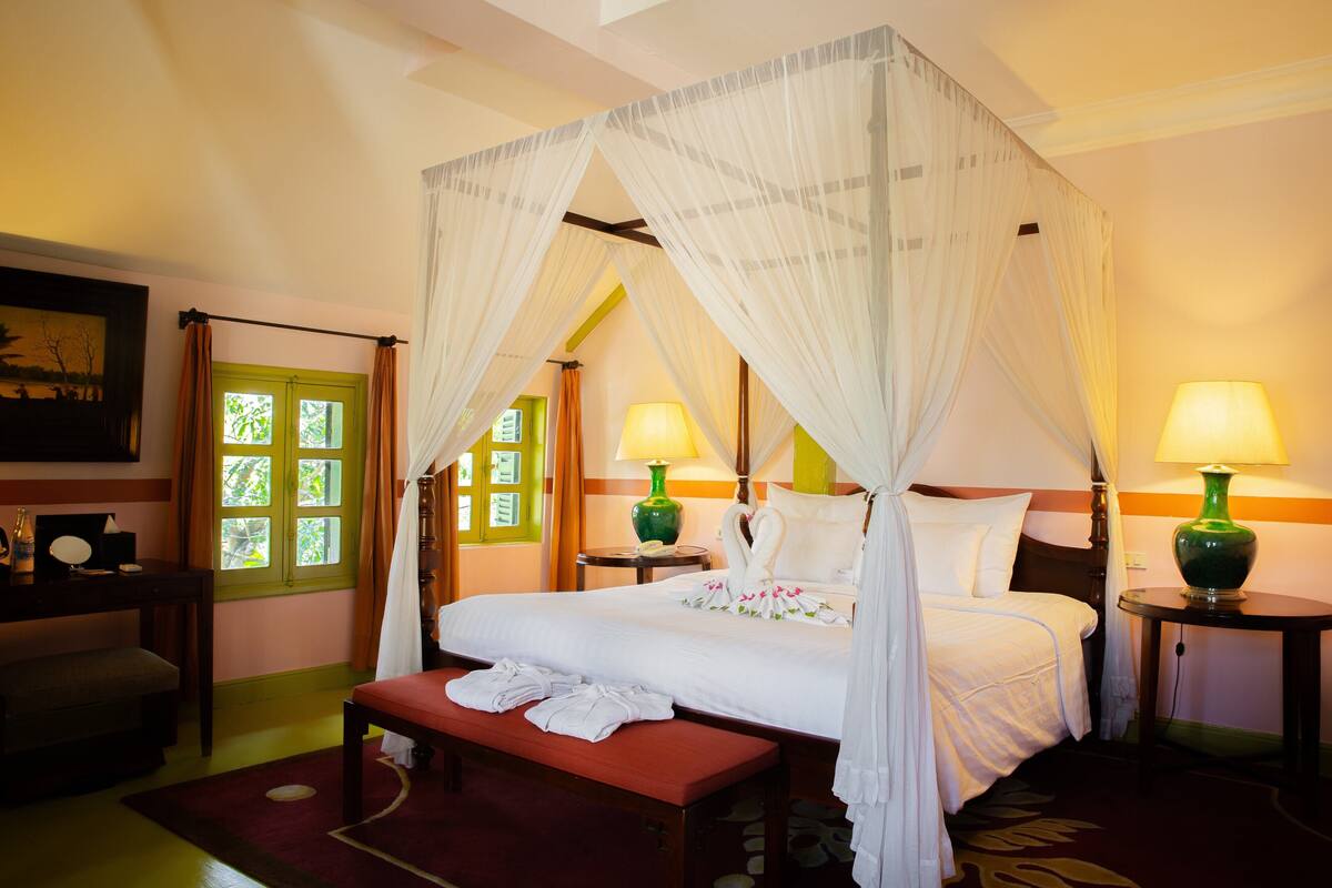 Una habitación de hotel con una cama con dosel, dos lámparas de noche, un banco con toallas y un ventanal con marco verde.
