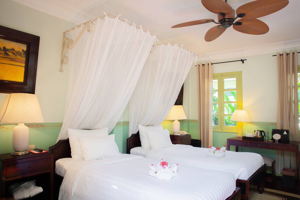 Una habitación de hotel con dos camas, un ventilador de techo, una mesita de noche con lámpara y una ventana con cortinas.