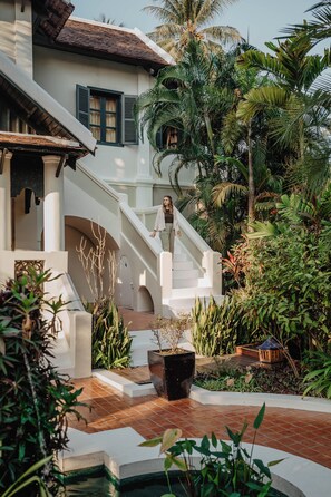 Ein weißes Gebäude mit einer Treppe, die zu einem Balkon führt, umgeben von üppiger Vegetation und einem kleinen Pool.