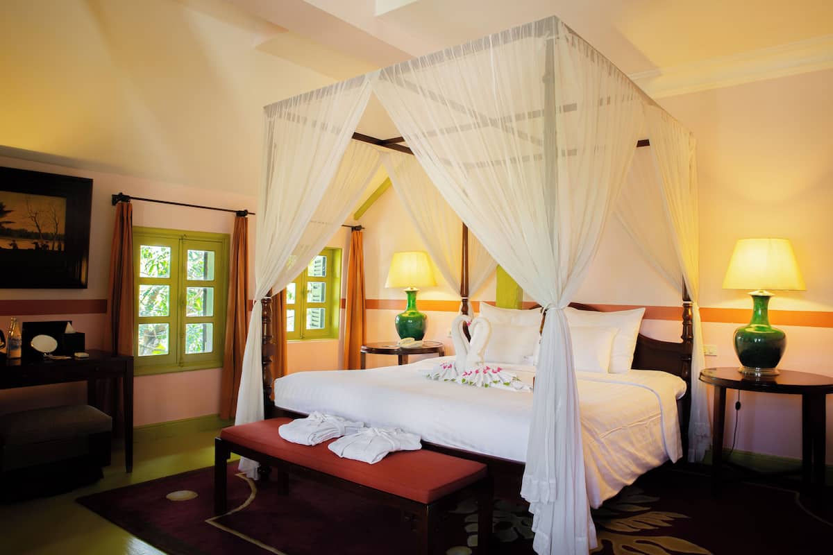 A four-poster bed with a canopy, bedside lamps, a bench with towels, and a desk with a chair.