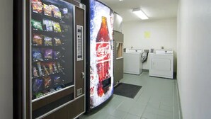 Dans une pièce au sol carrelé et aux murs blancs, on trouve un distributeur automatique de collations, une publicité pour Coca-Cola, un lave-linge et un sèche-linge.