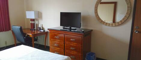 A hotel room with a bed, a desk with a computer, a dresser, a mirror, and a trash can.