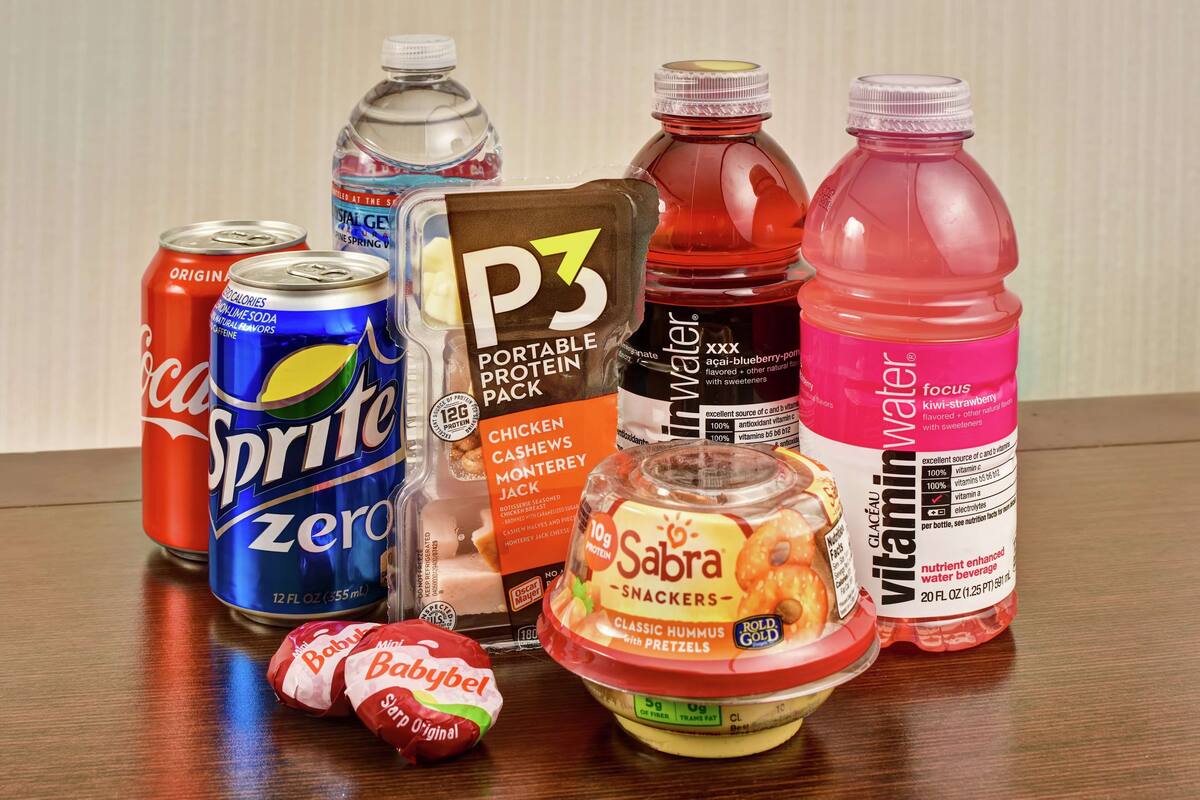 A variety of food and drink items including a can of Sprite Zero, a portable protein pack, a bottle of water, a bottle of vitamin water, a container of Sabra snacks, and a Babybel candy.