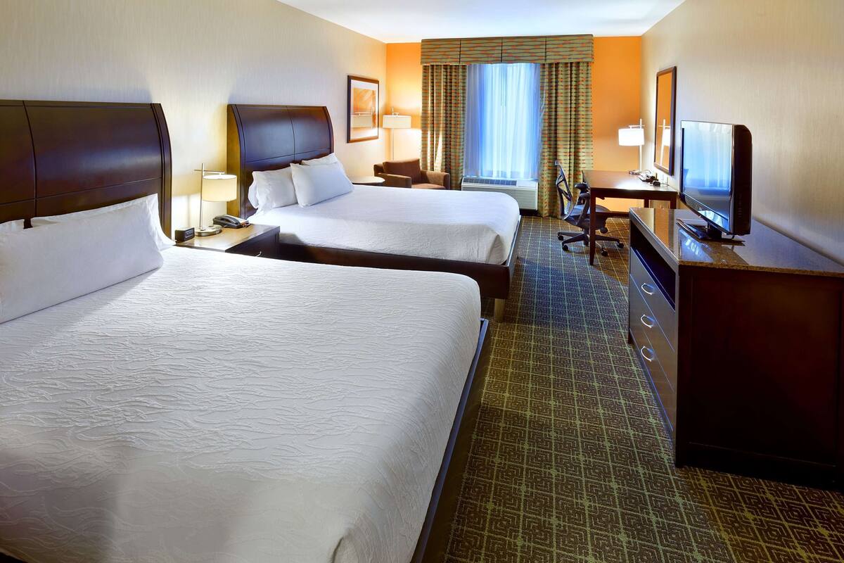 A hotel room with two beds, a desk, a television, and a window with curtains.
