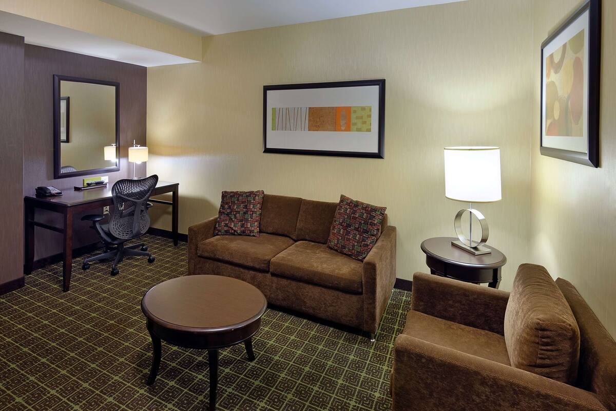 A hotel room with a brown sofa, a matching armchair, a small round table, a desk with a chair, a lamp, and a framed abstract painting on the wall.