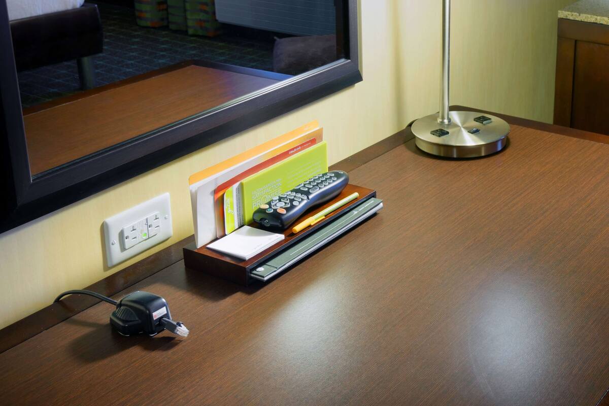 A hotel room desk with a lamp, remote controls, a power strip, and a window with a view of the hotel interior.