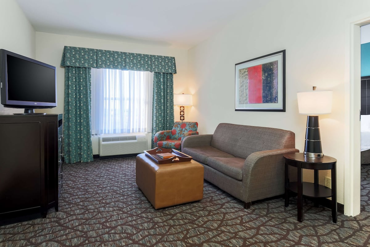 A hotel room with a sofa, ottoman, television, and a framed picture on the wall.