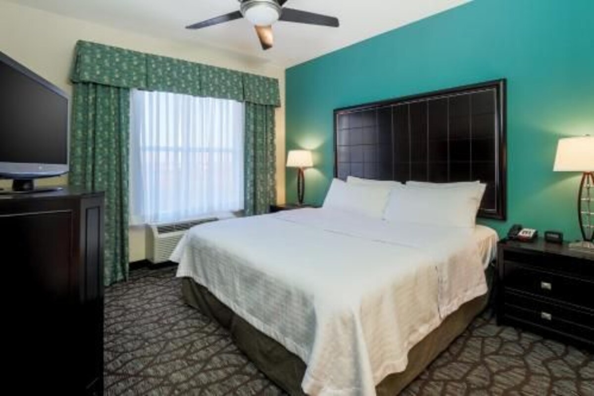 Habitación de hotel con cama, televisión, ventilador de techo, ventana con cortinas y mesita de noche con lámpara.