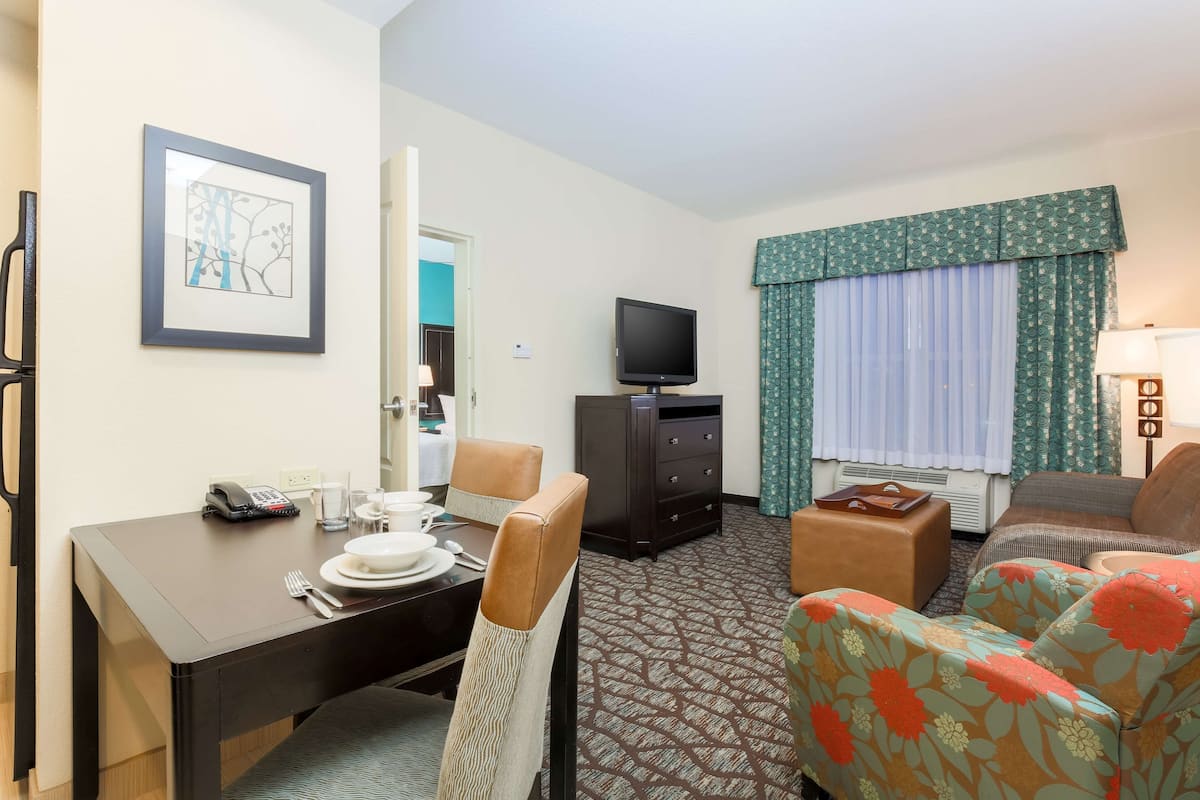 A hotel room with a dining table set for two, a television, a sofa, and a patterned carpet.