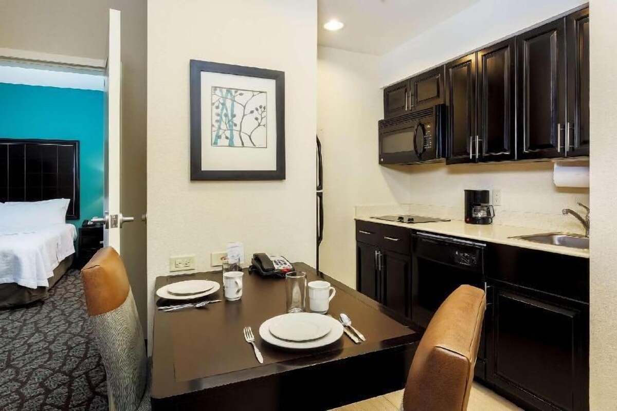 Habitación de hotel con cama, una pequeña mesa de comedor con platos, tazas y cubiertos, un microondas y una cafetera en la zona de la cocina.