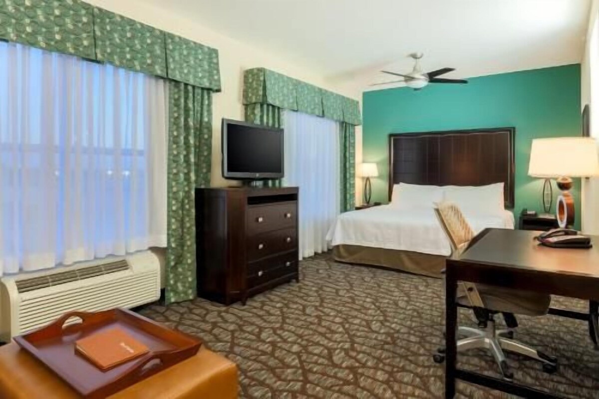 Habitación de hotel con cama, escritorio, televisor, ventilador de techo y ventana con cortinas.