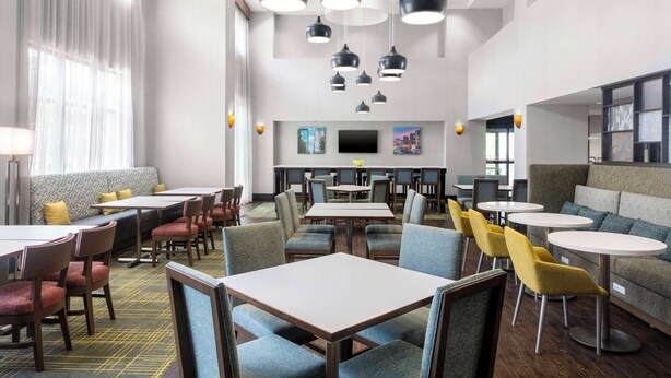 A modern dining area with multiple tables and chairs, a sofa, and a television on the wall. - Hampton Inn & Suites Birmingham/280 East-Eagle Point (Birmingham)
