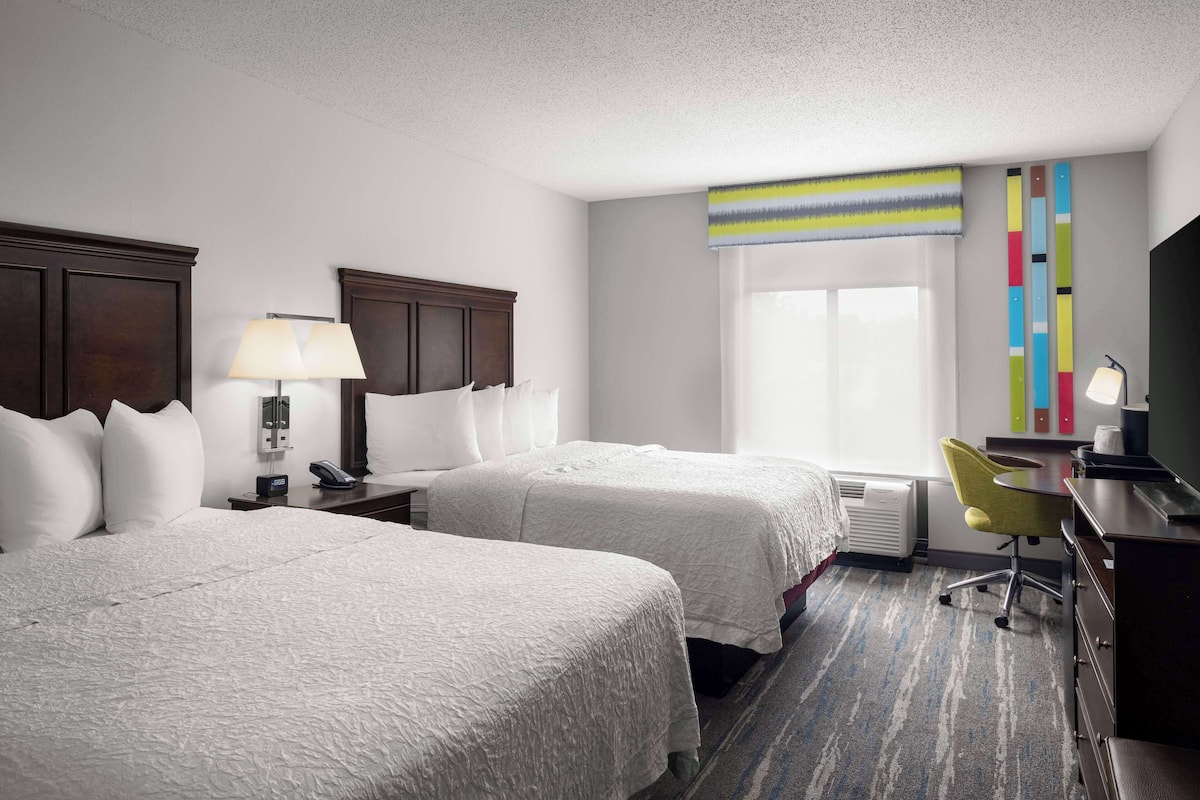 A hotel room with two beds, a desk, a chair, a lamp, and a window with a view.