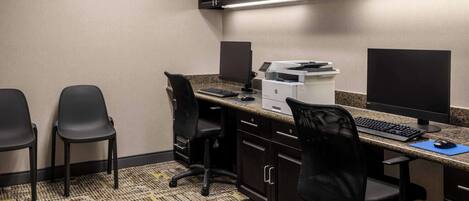 A hotel office with two black chairs, a desk with a computer, printer, and a clock on the wall.