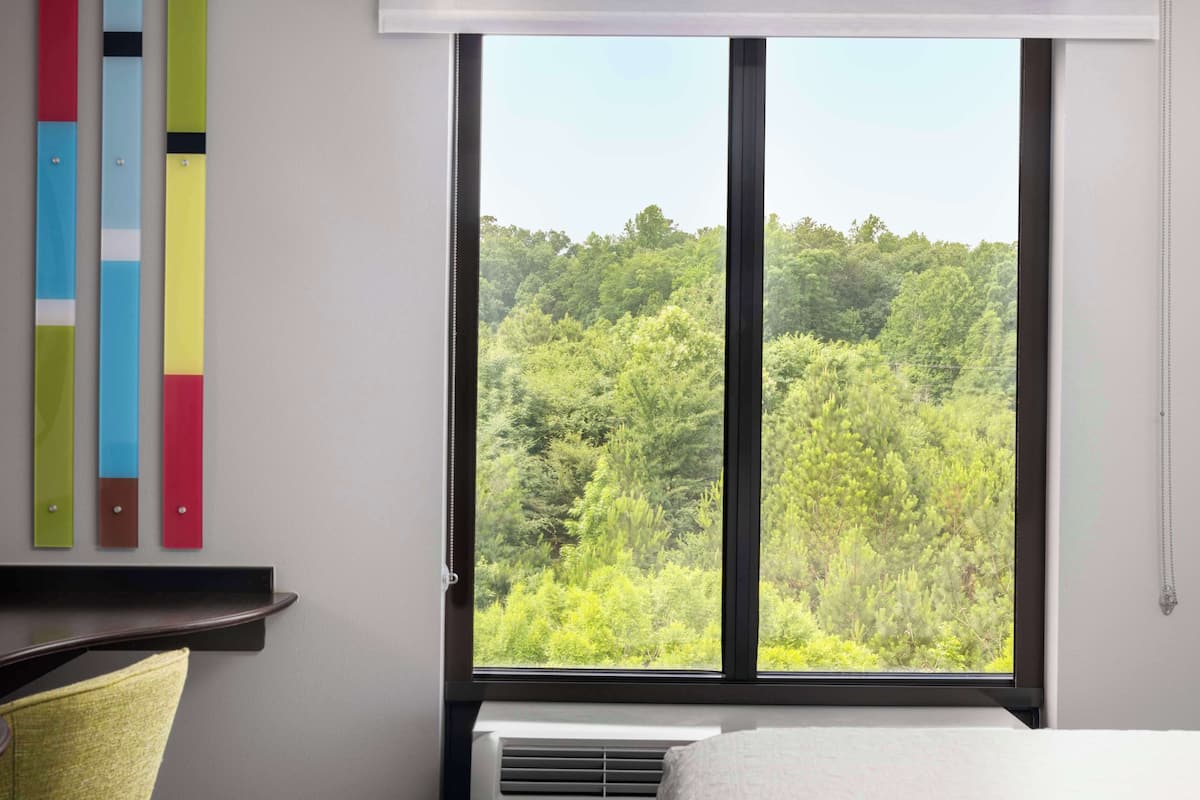 A room with a large window overlooking a forested area, a bed, a bedside table, and a wall-mounted shelf with colorful objects.