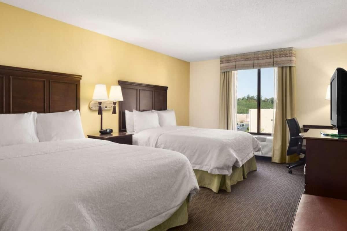 A hotel room with two beds, a desk, a TV, a window with curtains, and a lamp.