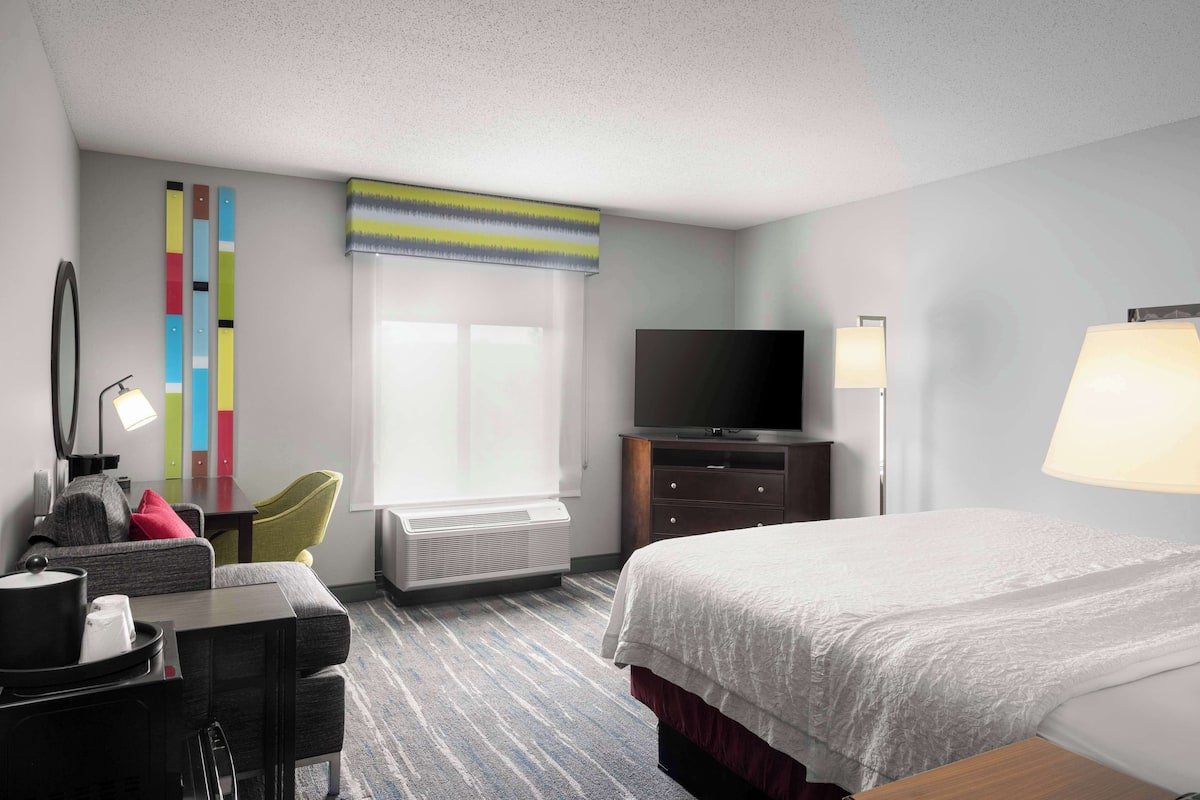 A hotel room with a bed, a TV, a desk with a lamp, a chair, and a window with blinds.