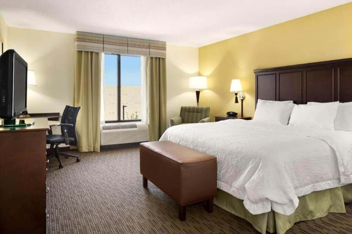 A hotel room with a bed, a desk with a computer, a chair, a lamp, and a window with curtains.