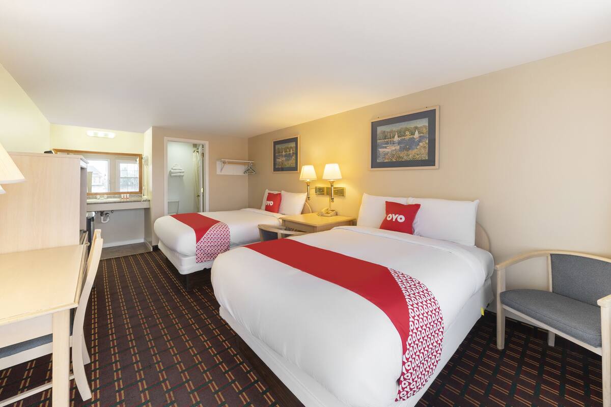 A hotel room with two beds, a desk, a chair, a small table, and a bathroom visible through an open door.