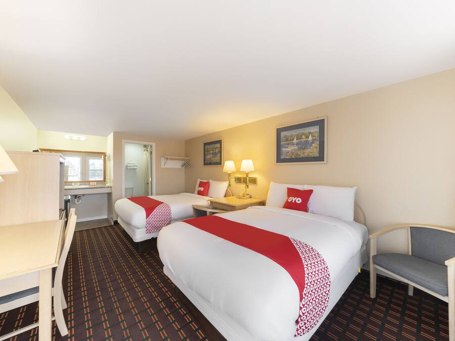 A hotel room with two beds, a desk, a chair, a small table, and a bathroom visible through an open door.