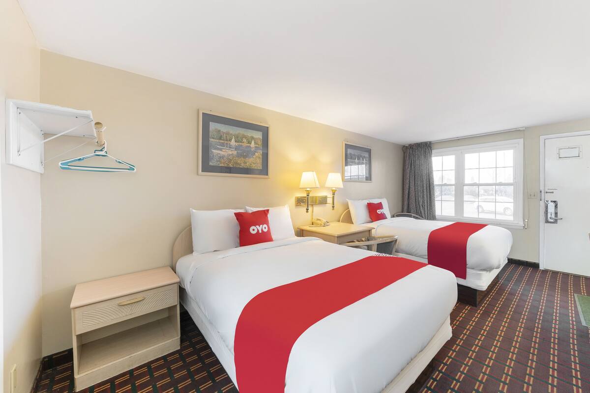 A hotel room with two beds, a desk, a chair, a hanging coat rack, and a window with curtains.